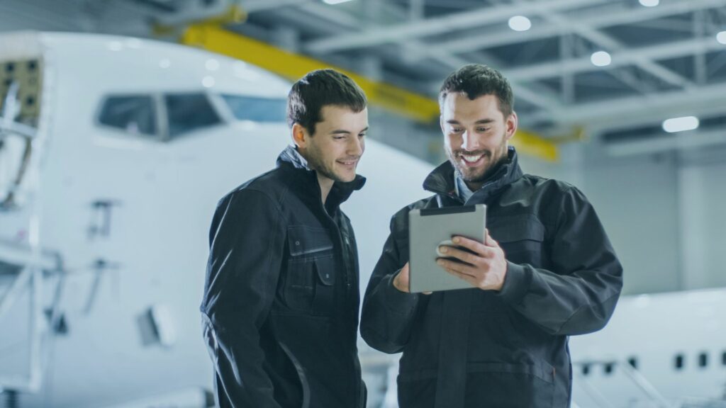 Two men are looking at an Ipad with airplanes in the background.